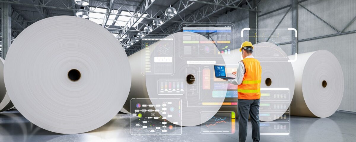 Worker in an orange safety vest using a laptop in a warehouse with large paper rolls and digital interface displays.