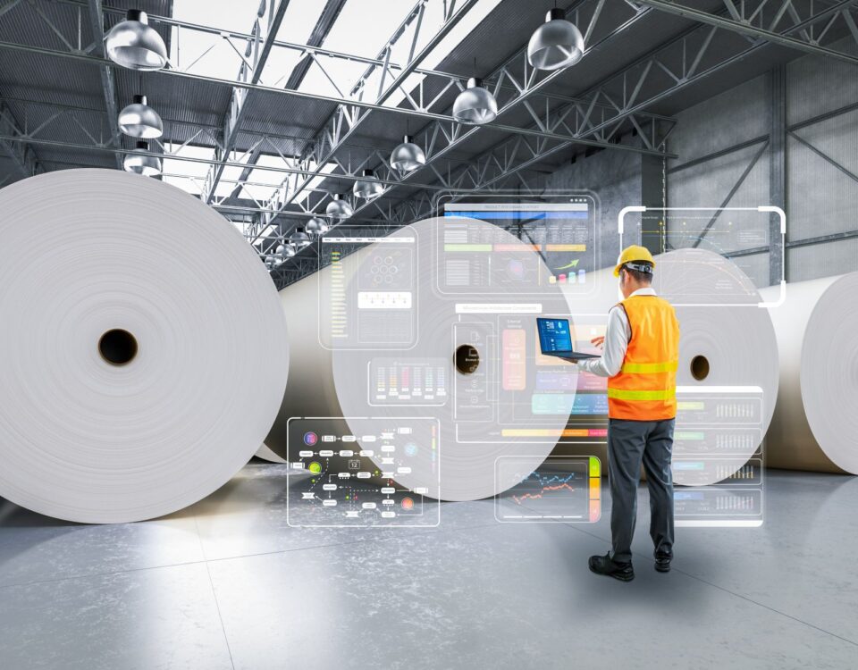 Worker in an orange safety vest using a laptop in a warehouse with large paper rolls and digital interface displays.