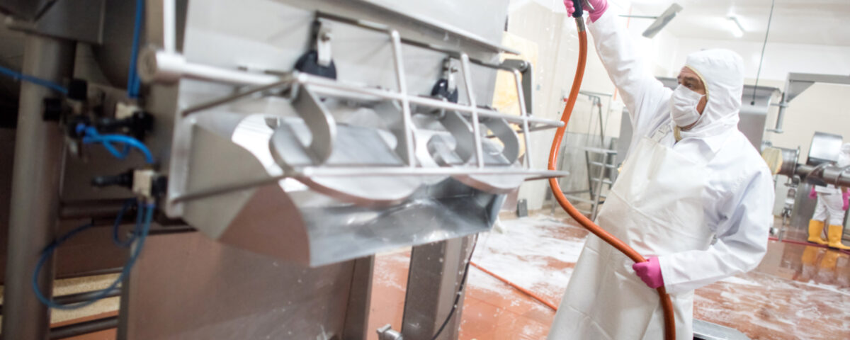 Worker in protective gear using a hose to clean industrial machinery in a processing facility.