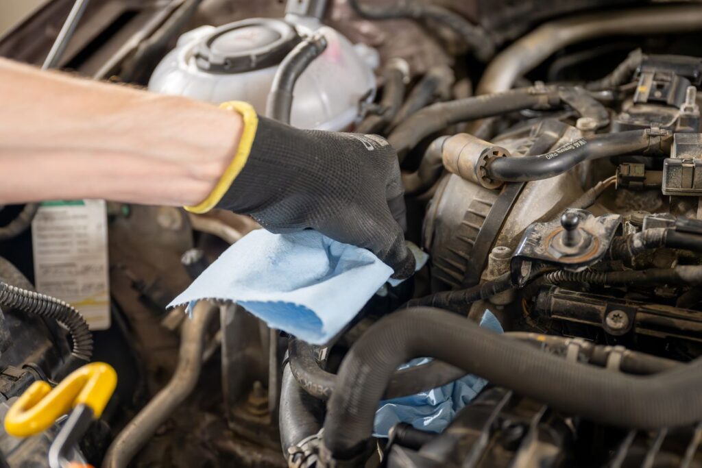 A mechanic wearing a black glove uses a blue cloth to clean engine components in a vehicle.