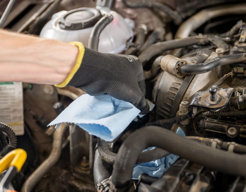 A mechanic wearing a black glove uses a blue cloth to clean engine components in a vehicle.