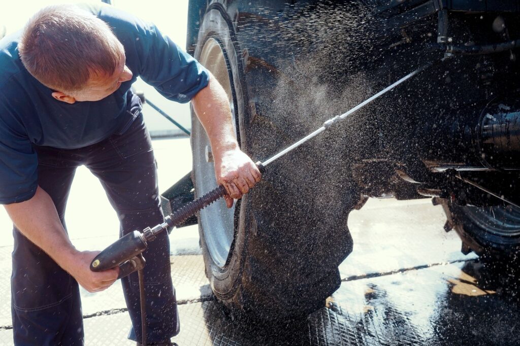 Individual operating a pressure washer to clean the wheels and body of a tractor.
