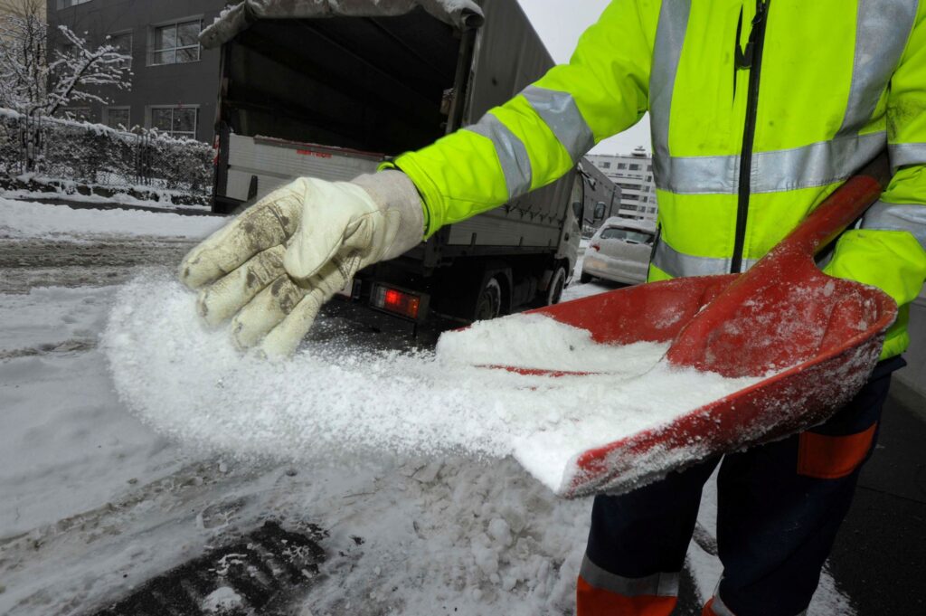 Individual in a high-visibility jacket using a shovel to spread salt on a snowy surface.