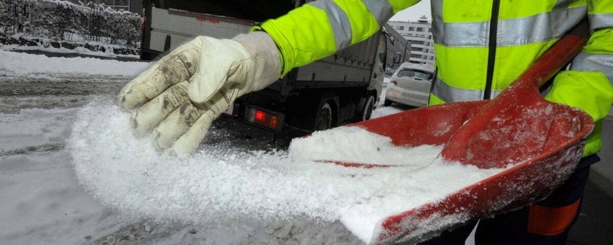 Individual in a high-visibility jacket using a shovel to spread salt on a snowy surface.