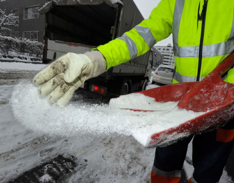 Individual in a high-visibility jacket using a shovel to spread salt on a snowy surface.