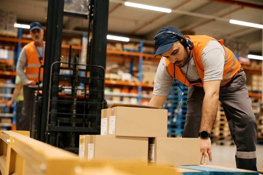 Warehouse worker in an orange vest lifting cardboard boxes while wearing headphones in a storage facility.