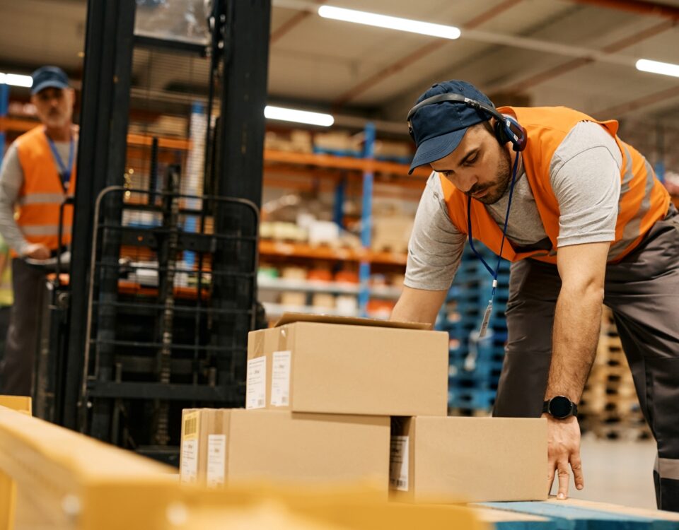 Warehouse worker in an orange vest lifting cardboard boxes while wearing headphones in a storage facility.